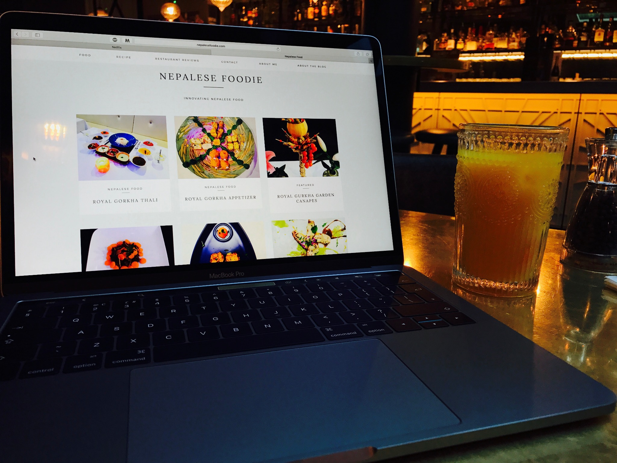 A laptop at a restaurant table showing the earlier Nepalese Foodie website beside an amber drink.